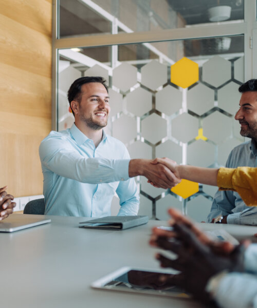 Group of professionals in a meeting room, with two people smiling and shaking hands across the table while others look on.