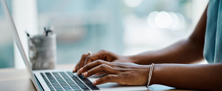 Close-up of a person typing on a laptop at a desk with a smartphone and papers nearby.